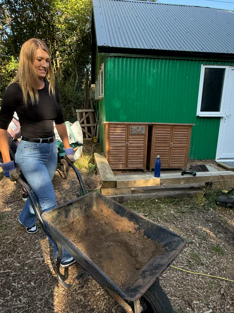 A volunteer pushing a wheelbarrow of soil past the green hut
