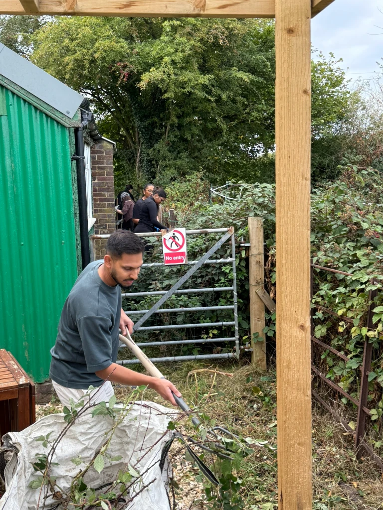 A volunteer forking cut brambles into a bag by the gate, with others working behind